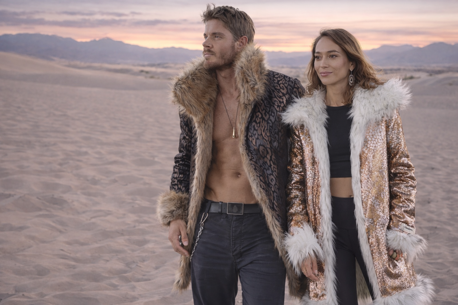 Two people wearing fur-lined jackets in a desert setting with a sunset.