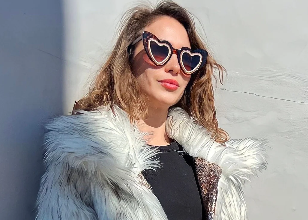 Woman wearing heart-shaped sunglasses and a white fur coat against a light background